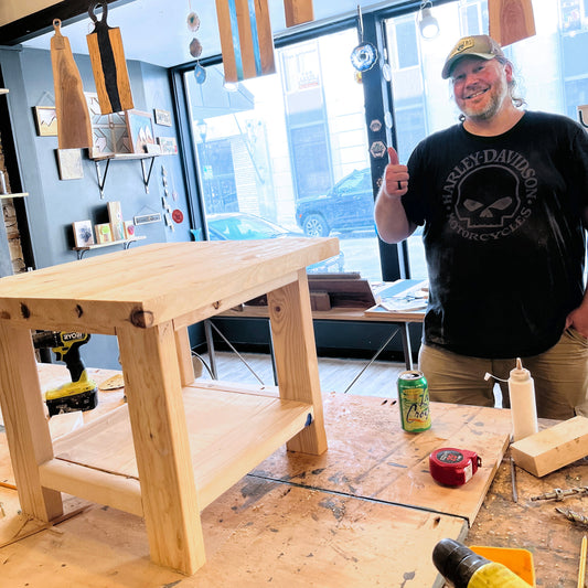 Man in a workshop with a wooden table and tools, wearing a black t-shirt with a graphic design.