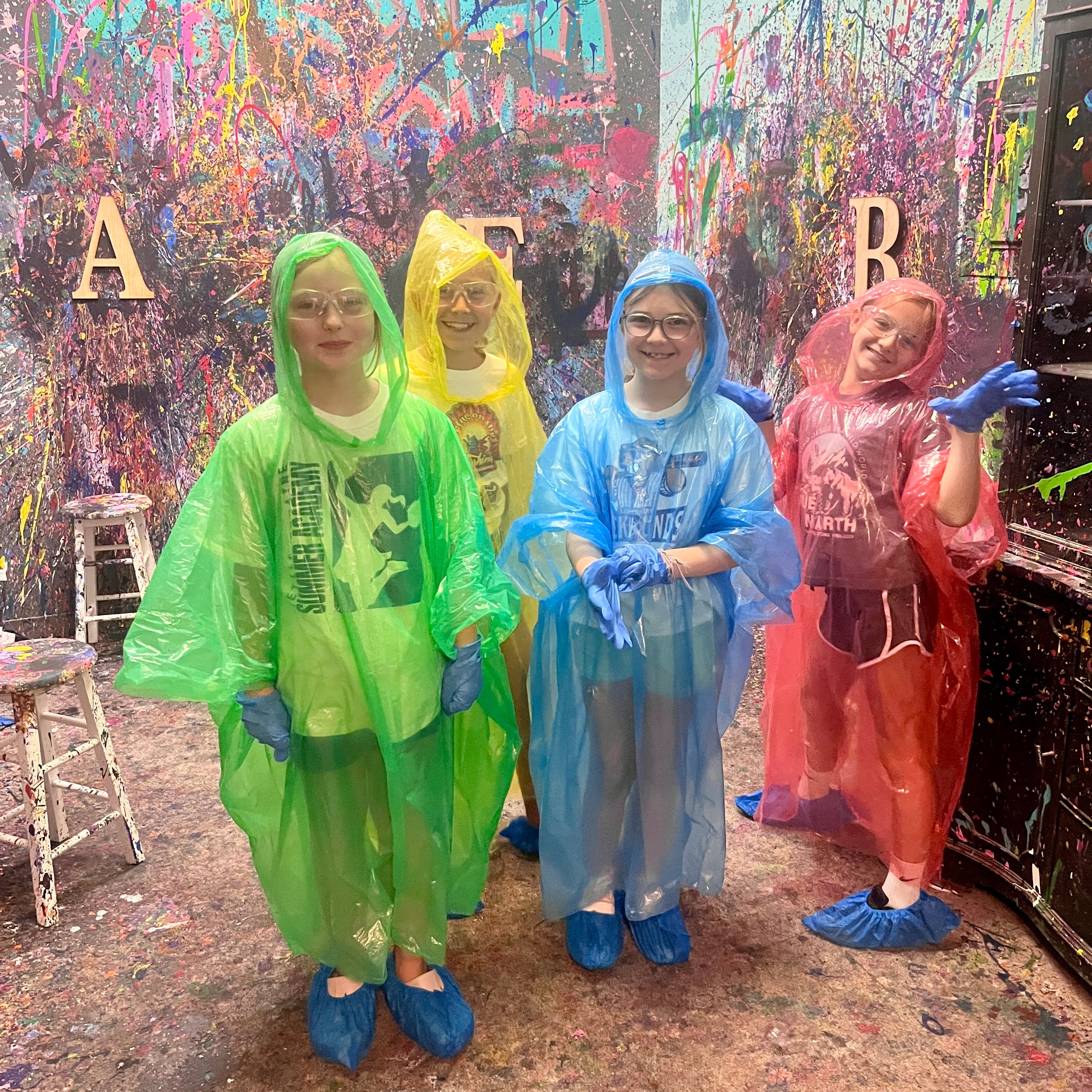 Four kids wearing colorful rain ponchos in front of a splatter room wall.