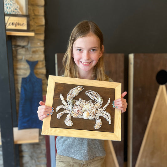 Young girl holding a framed string art piece of a a crab with shells in a studio setting.