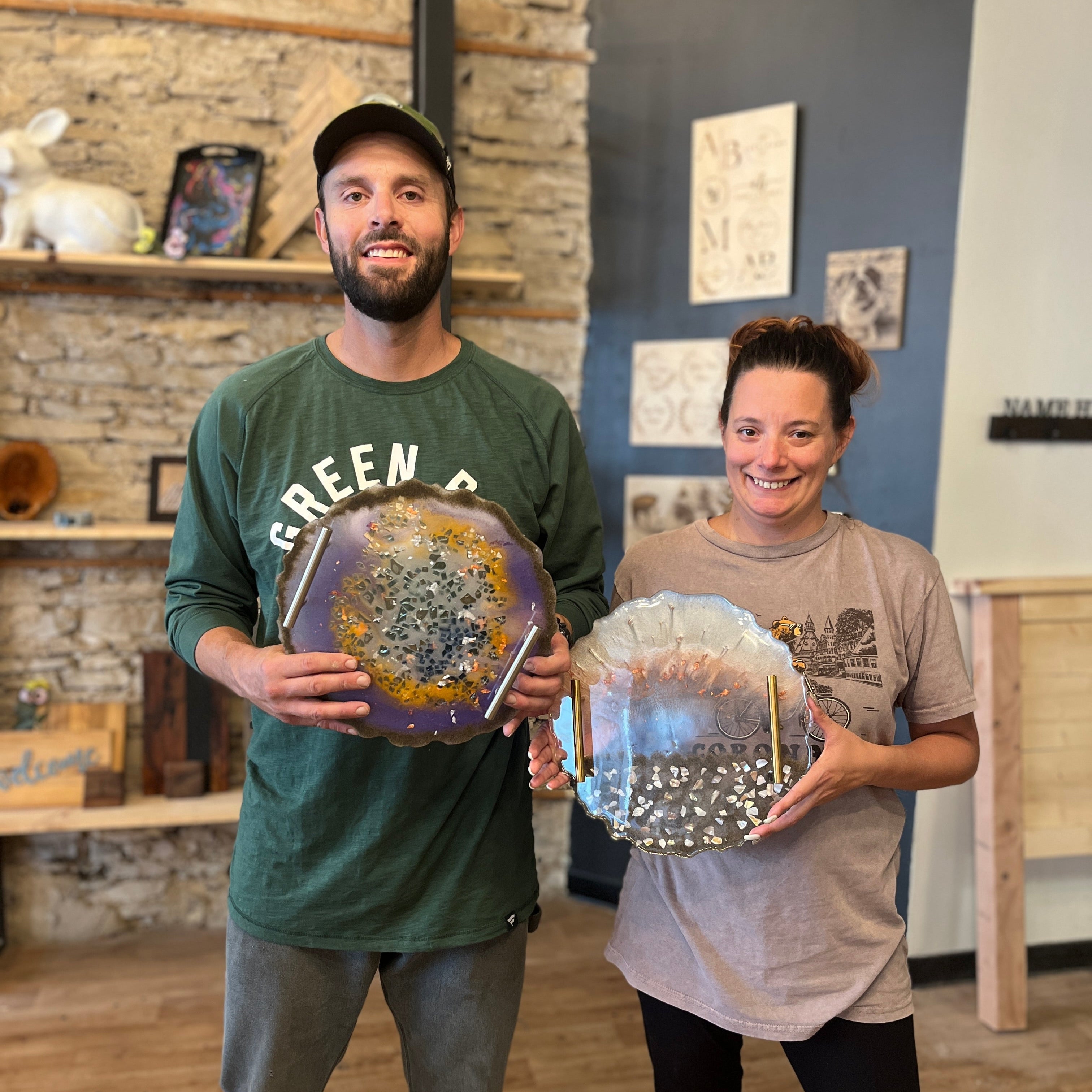Two people holding decorative epoxy geode serving trays in a room with a rustic wall and wooden floor.
