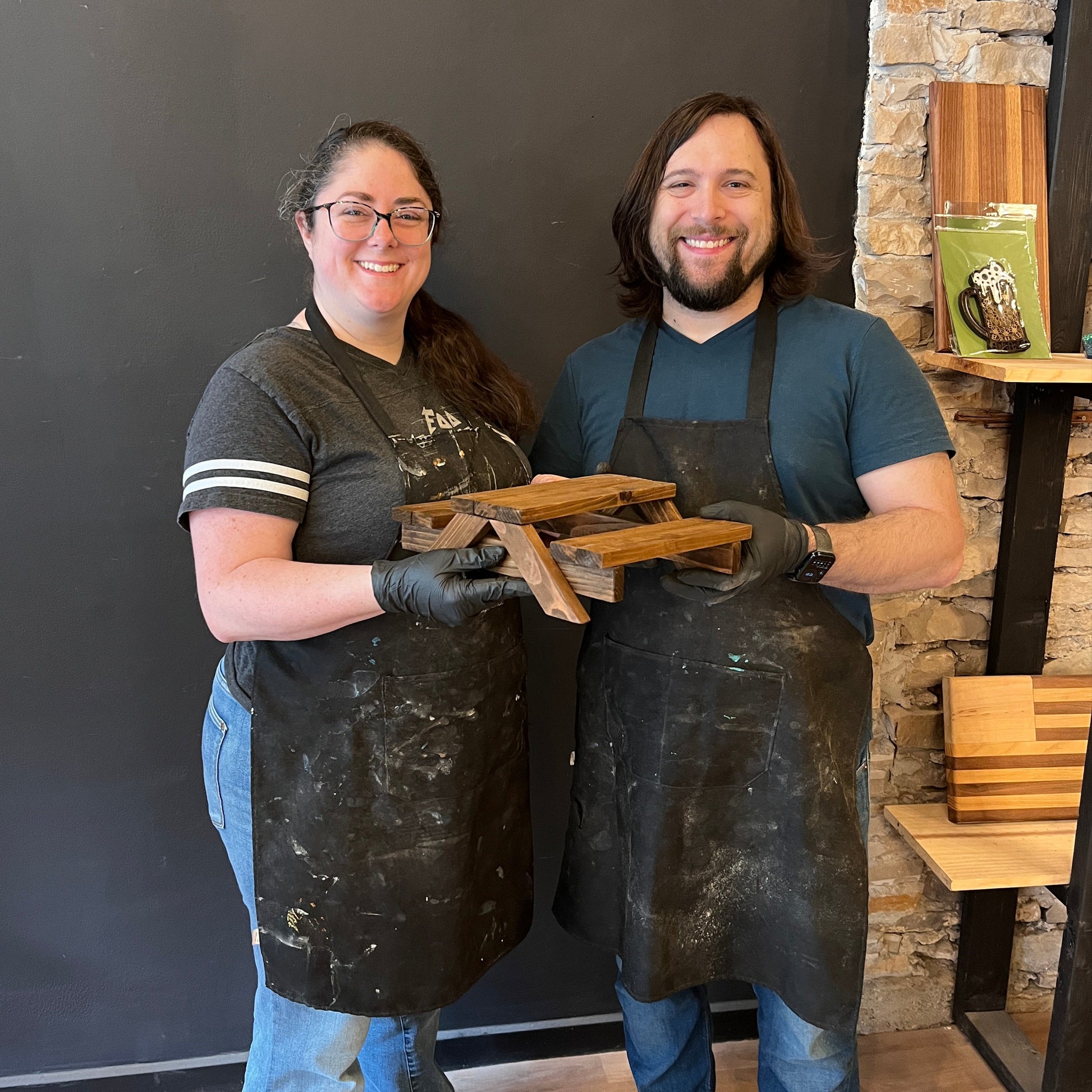 Two people wearing aprons and gloves, holding a mini picnic table, and standing in front of a stone wall.