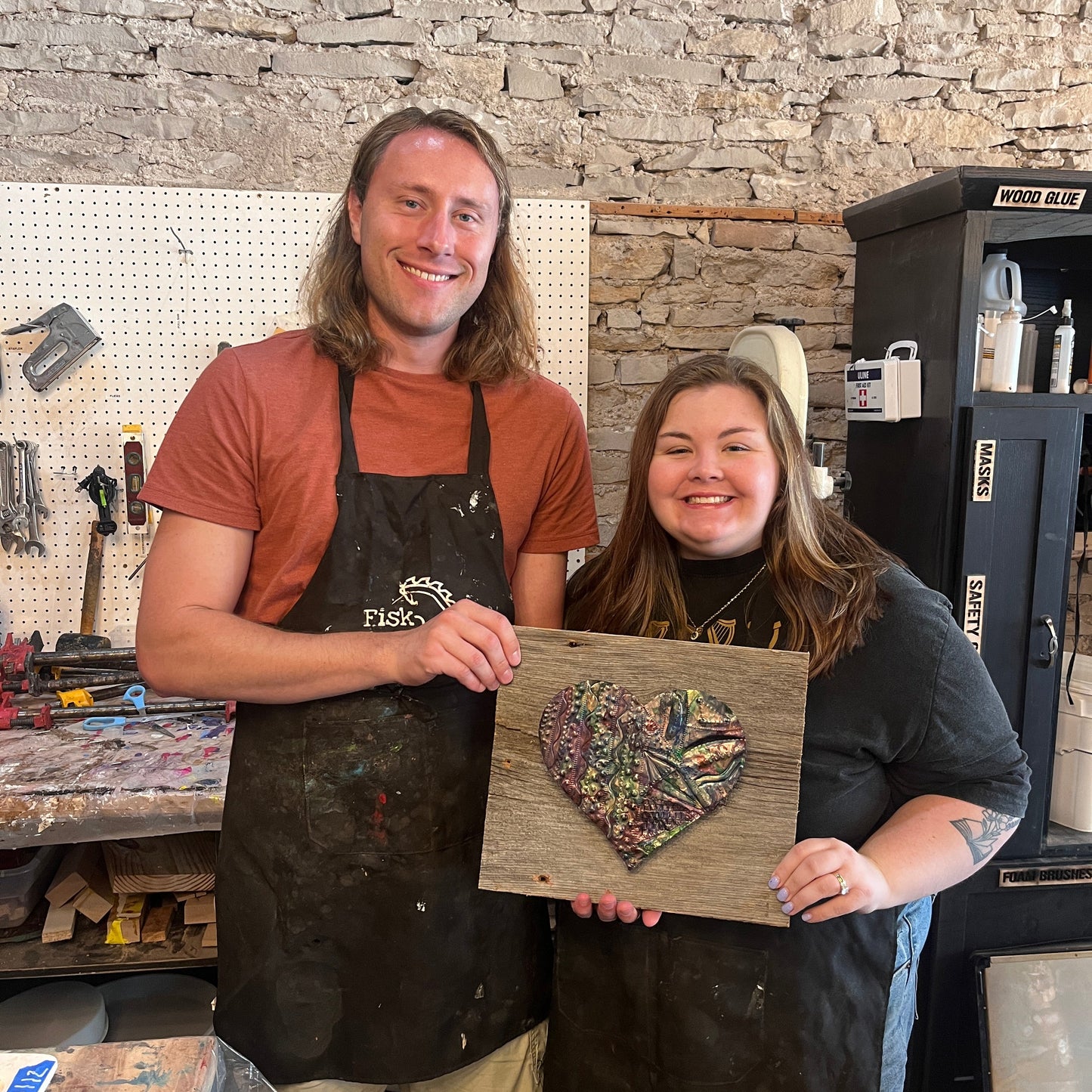 Two people holding a copper embossed heart on a wooden board, standing in a studio with a stone wall.