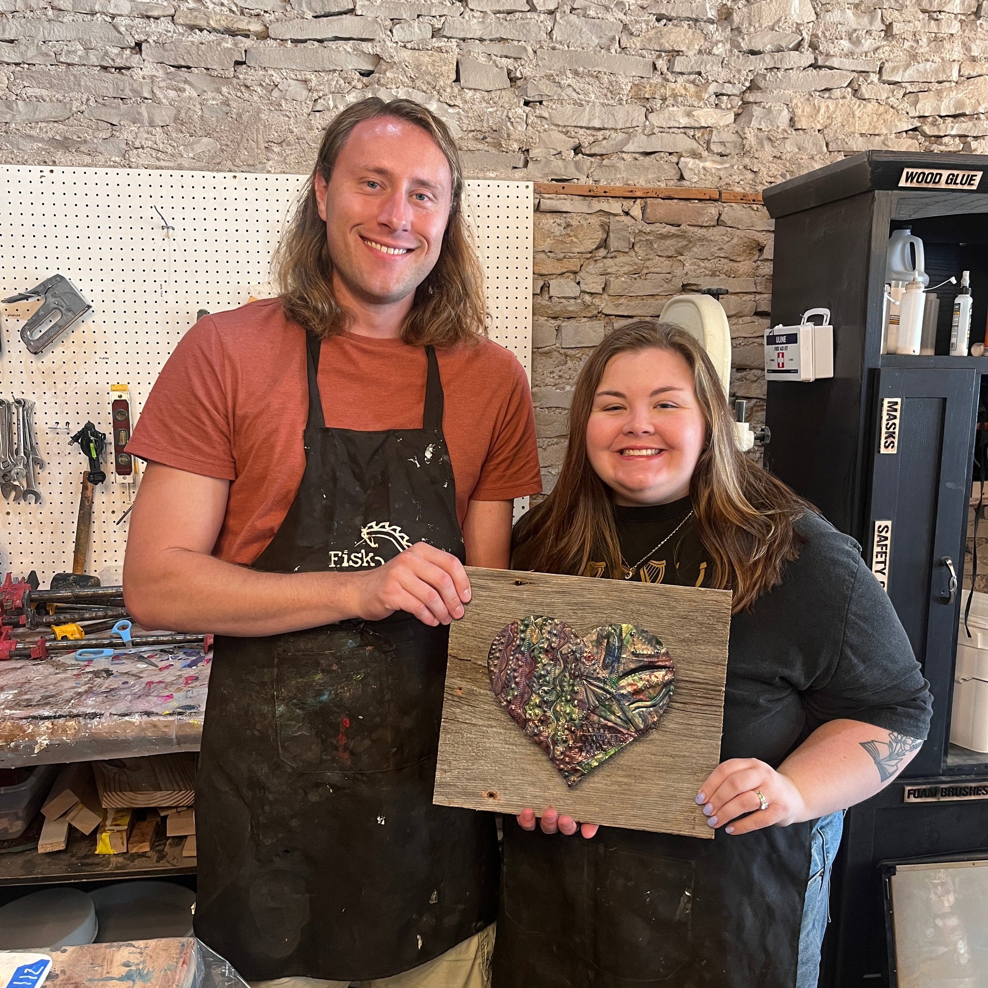 Two people holding a copper embossed heart on a wooden board, standing in a studio with a stone wall.