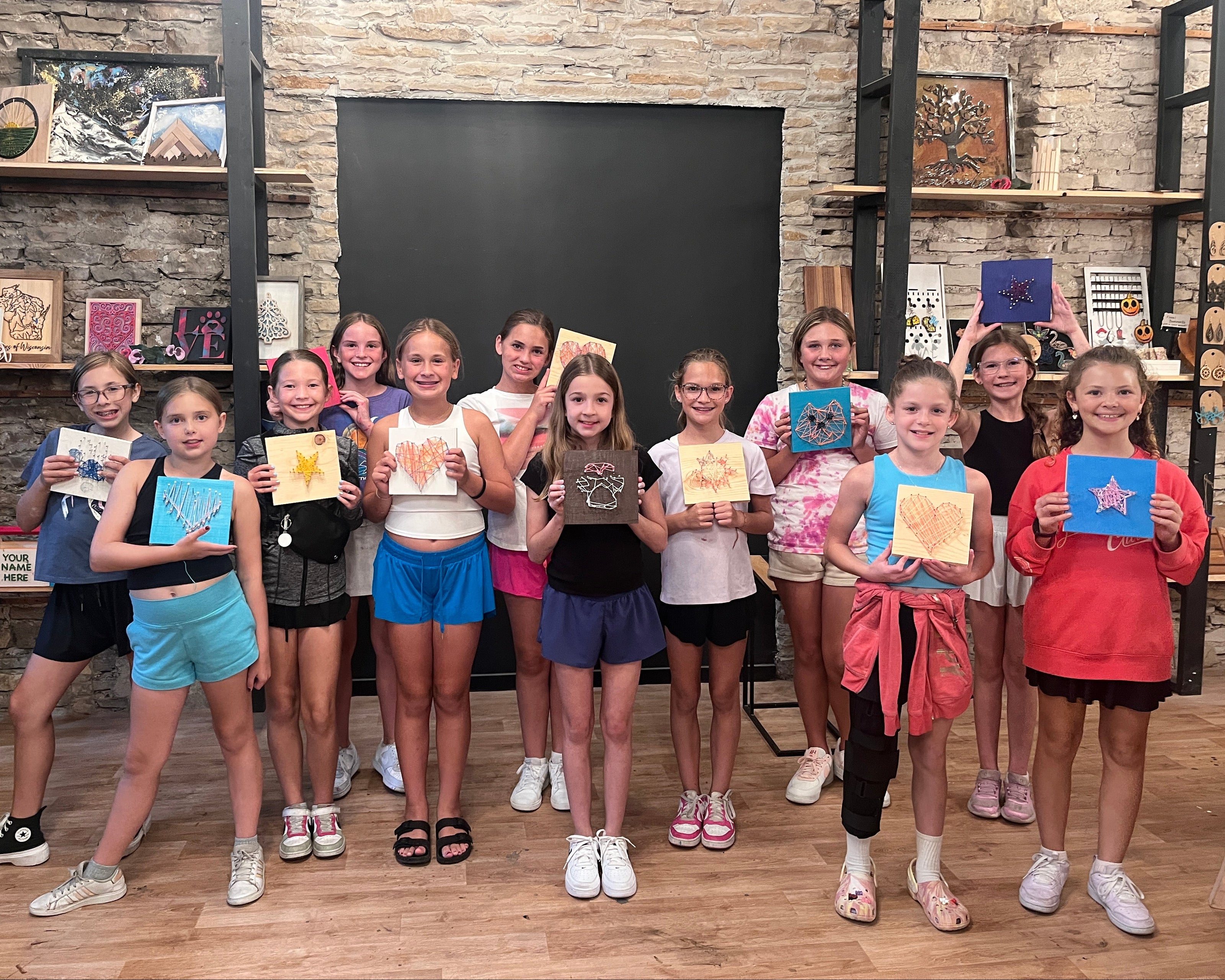 A group of girls at a birthday party holding up their string art in a studio with shelves and stone walls.