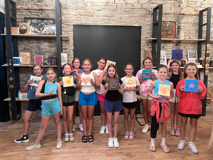 Group of children holding up their artwork in a room with shelves and a blackboard.