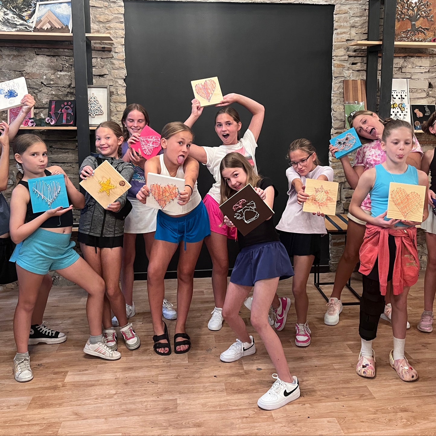 Group of girls holding up string art in a studio with brick walls and shelves.