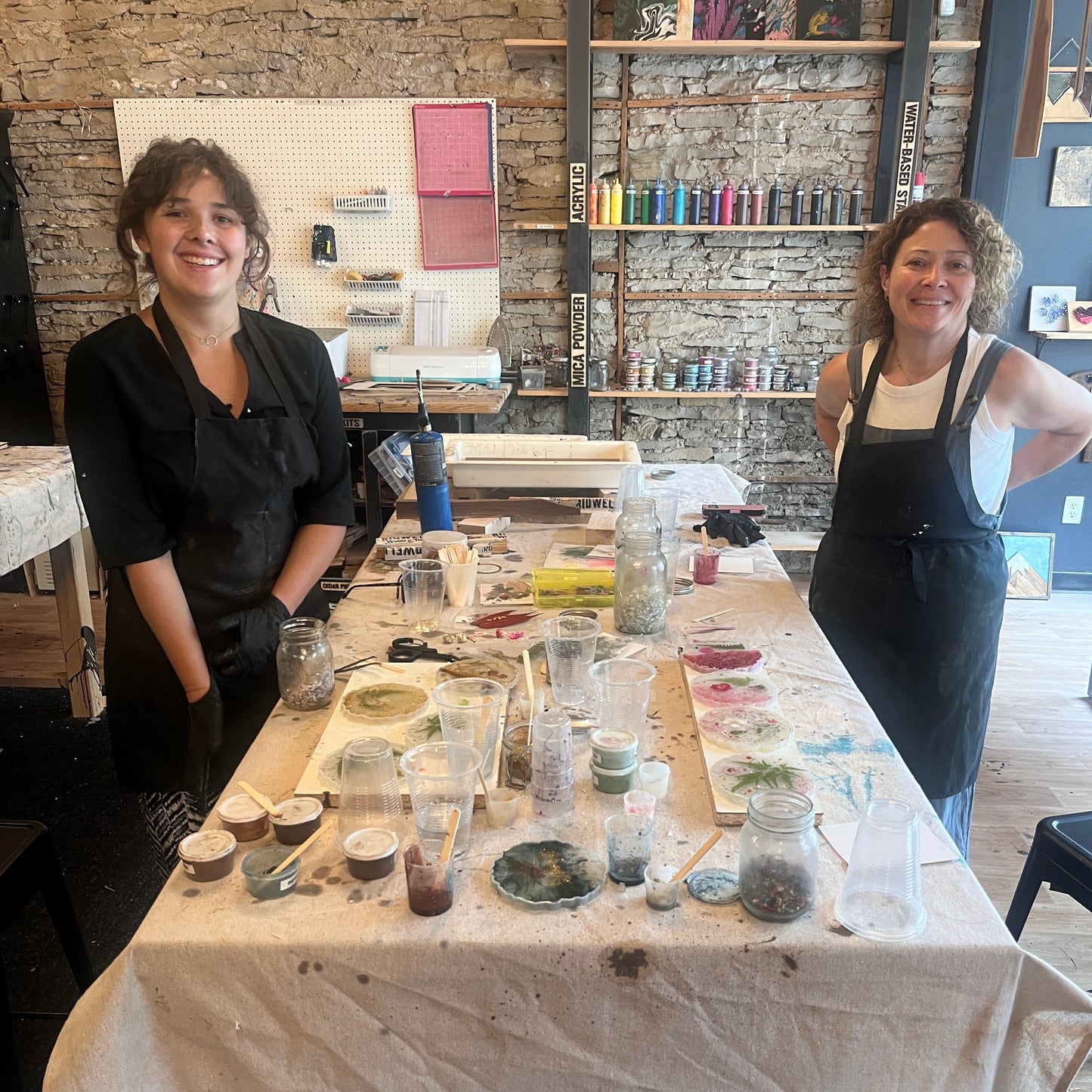 Women standing in front of a table with various epoxy coaster supplies in a workshop setting.