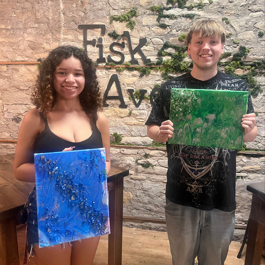 Two people holding up green and blue fluid art canvases against a stone wall.