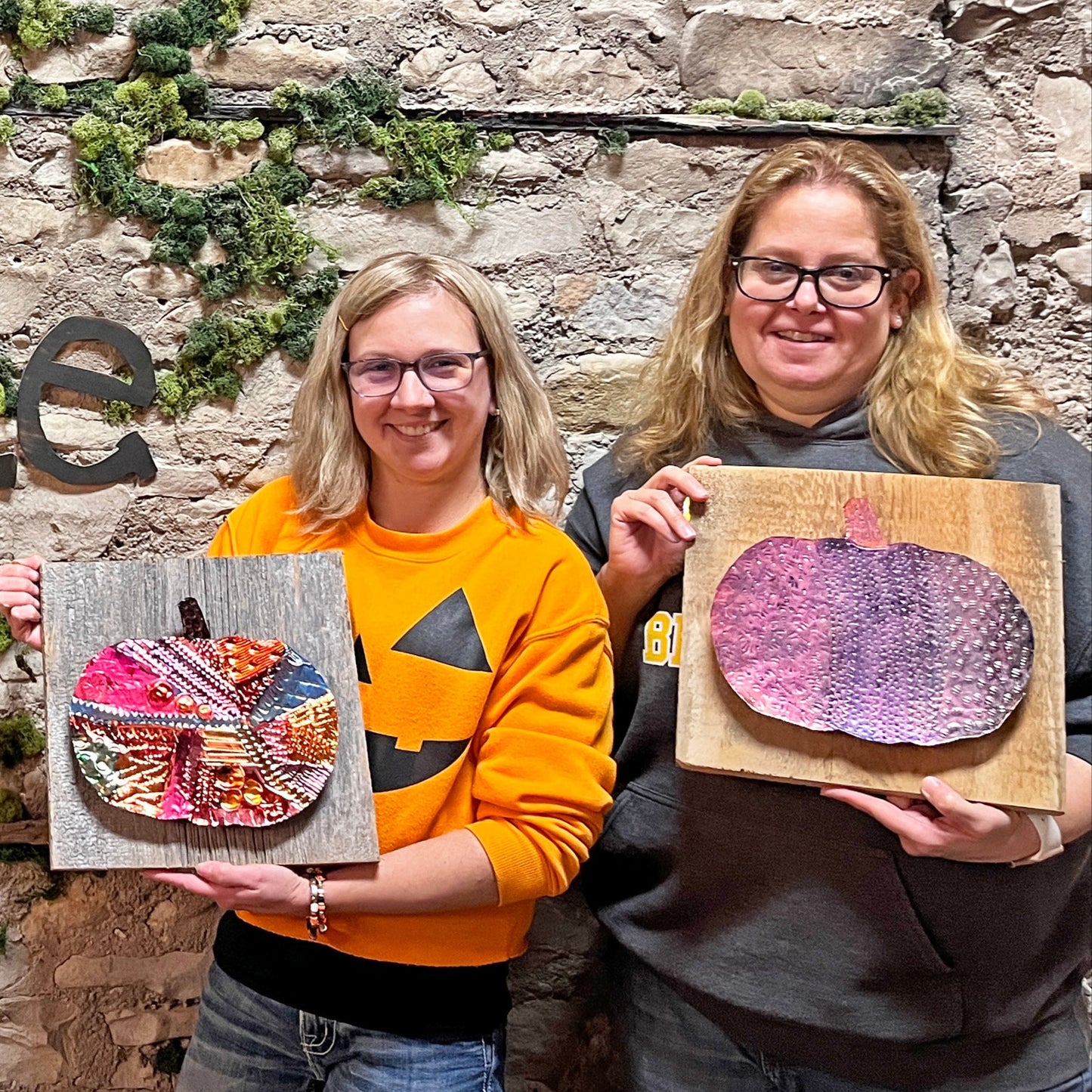 Two people holding copper pumpkin art in front of a stone wall with 'Fisk Avenue' sign.