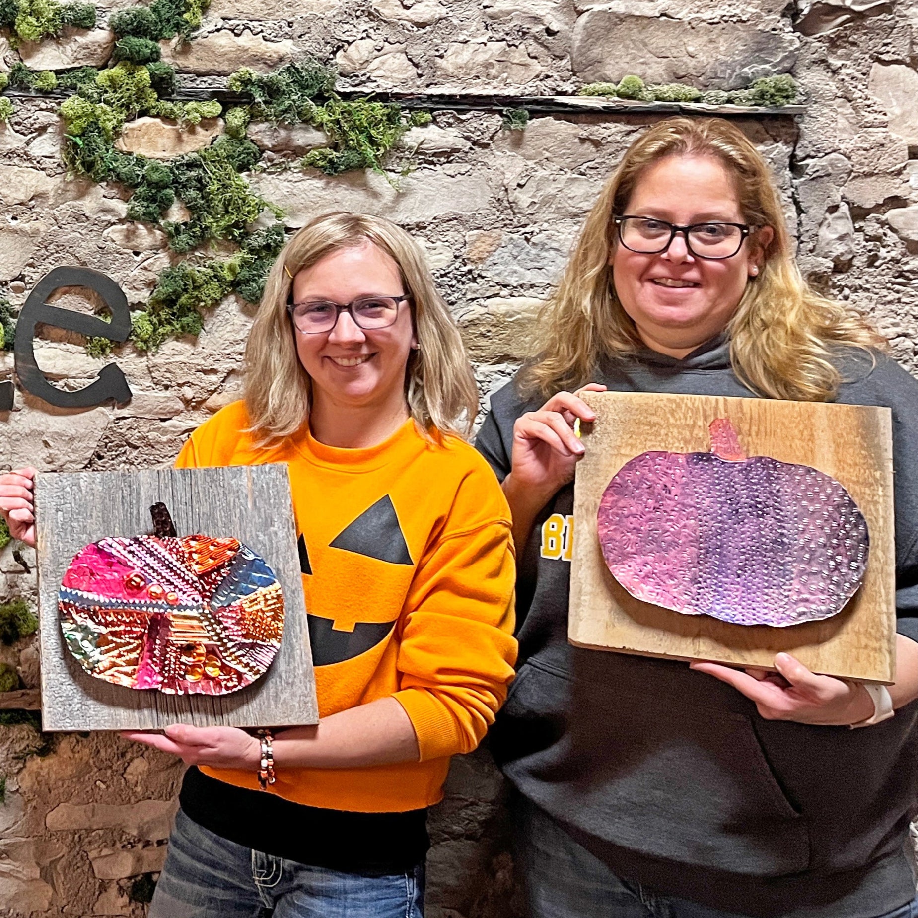 Two people holding copper pumpkin art in front of a stone wall with 'Fisk Avenue' sign.