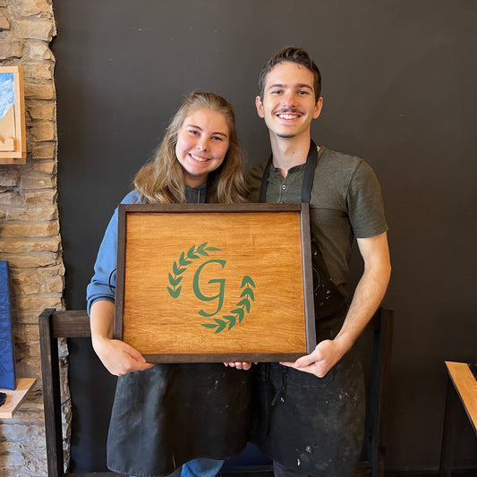 Two people holding a wooden serving tray with a logo in a rustic studio setting