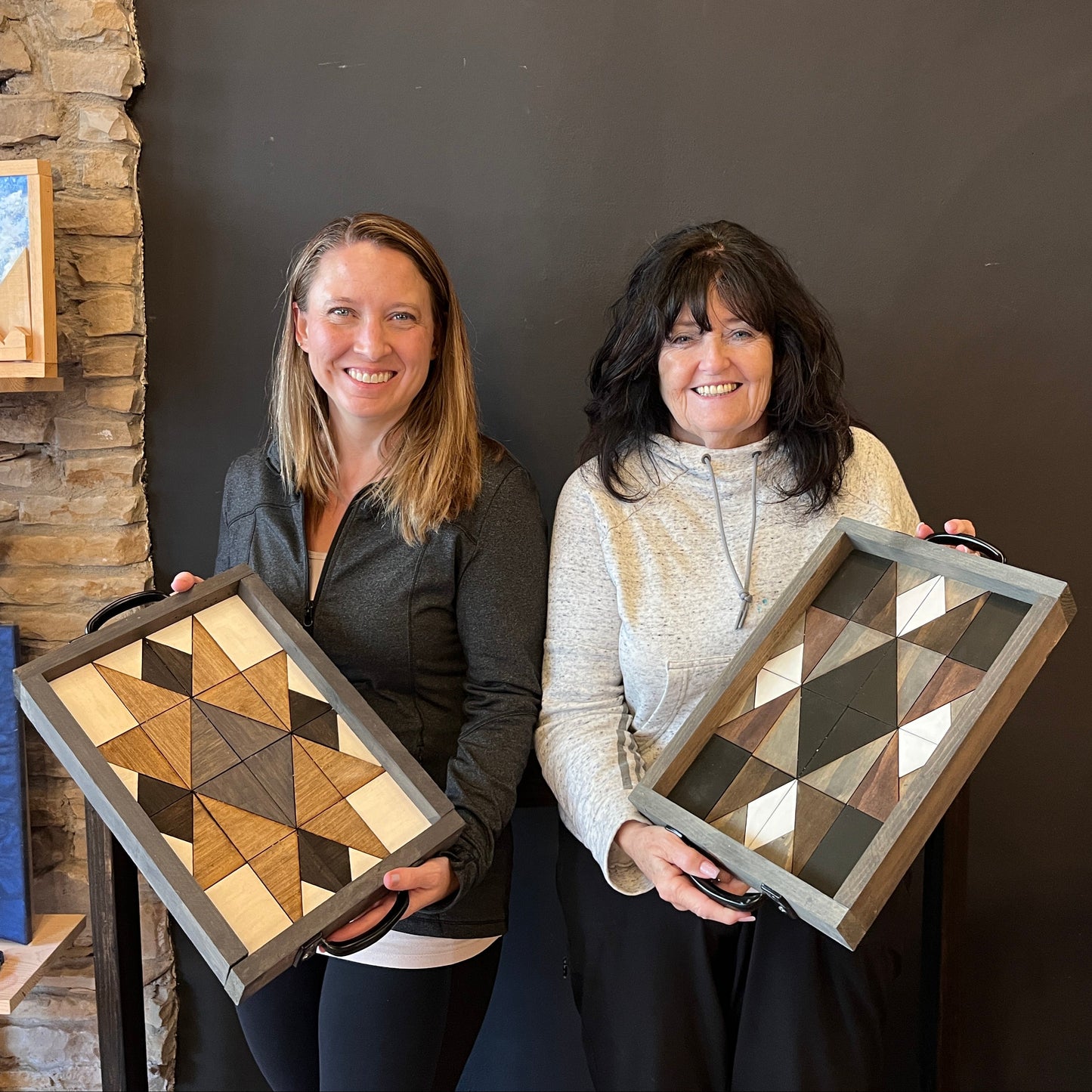 Two women holding wood mosaic geometric trays against a dark wall.