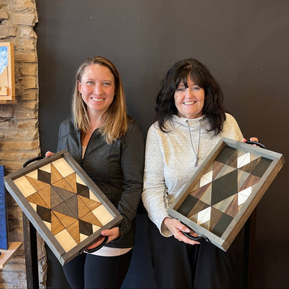 Two women holding wood mosaic geometric trays against a dark wall.