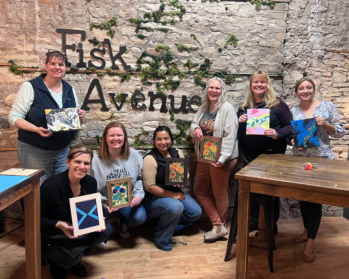 A group of seven women posing together in a studio with a rustic stone wall background that reads 'Fisk Avenue'. Each woman is holding a colorful, hand-painted framed artwork.
