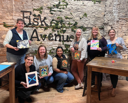 A group of seven women posing together in a studio with a rustic stone wall background that reads 'Fisk Avenue'. Each woman is holding a colorful, hand-painted framed artwork.
