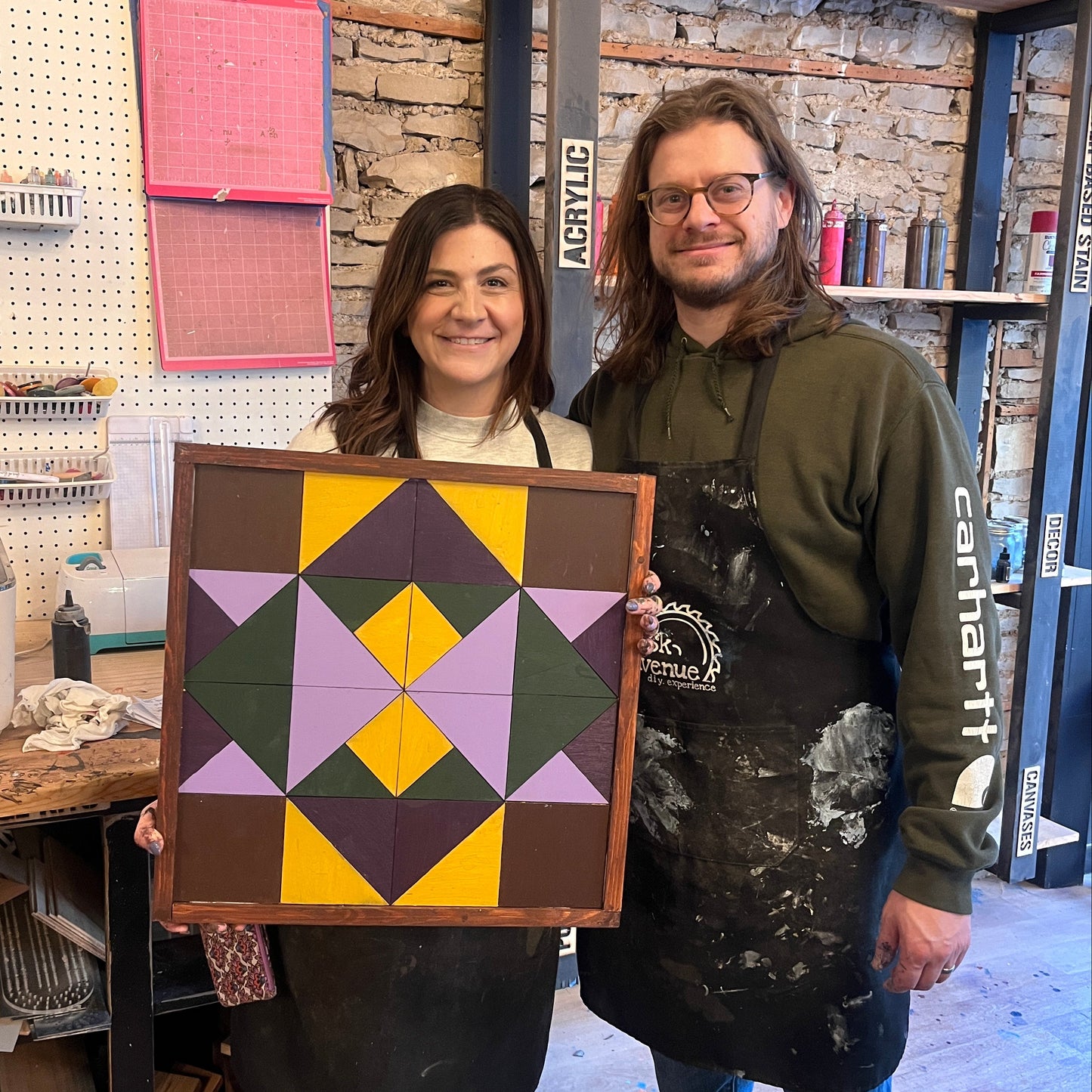 Two people holding a geometric patterned wooden barn quilt in a studio setting with tools and materials around