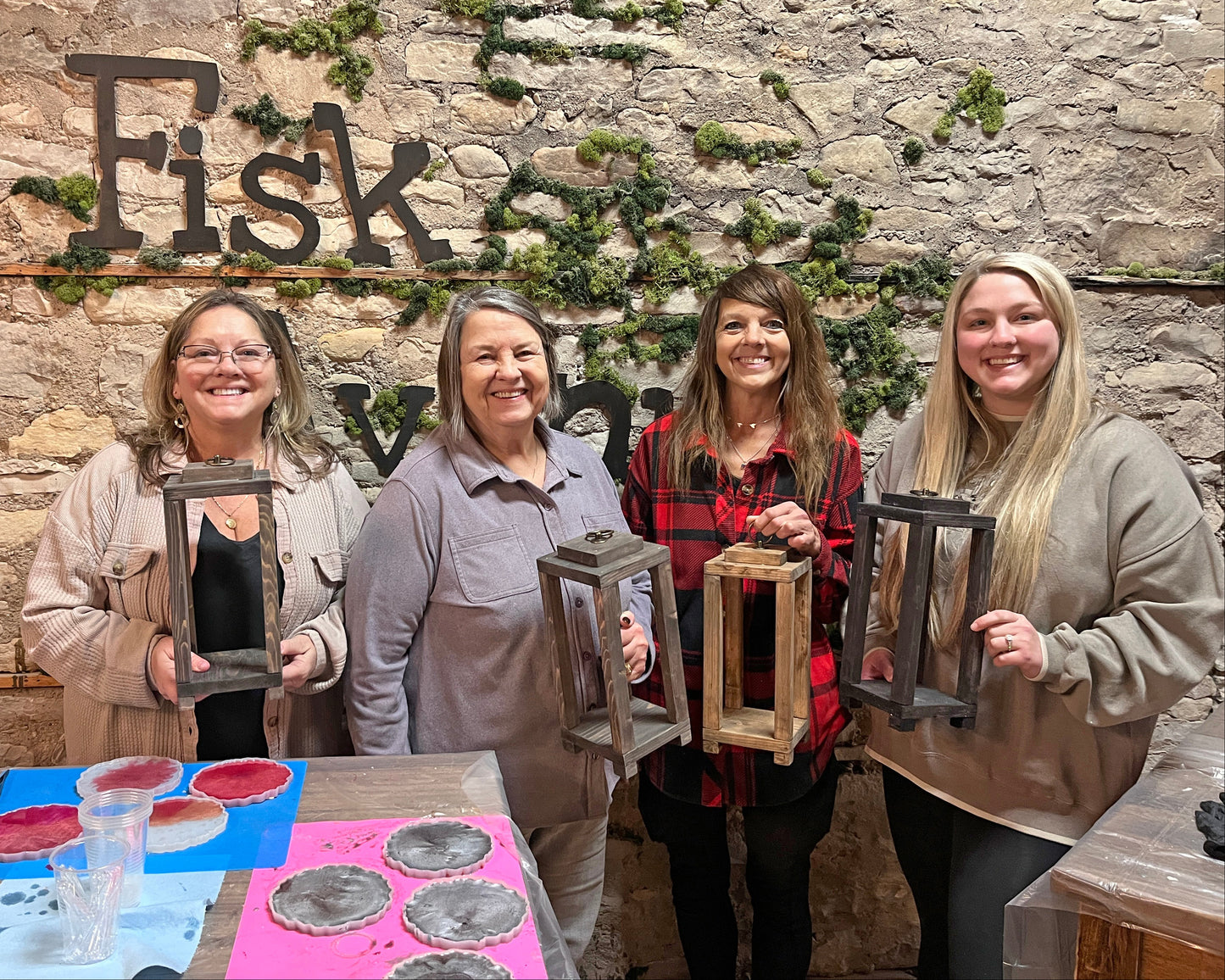 Four women holding wooden candle lanterns in front of a stone wall with 'Fisk Avenue' branding.