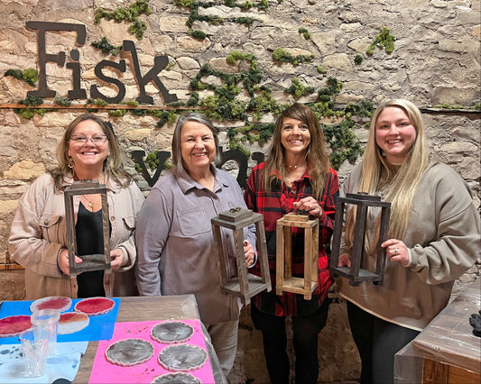 Four women holding wooden candle lanterns in front of a stone wall with 'Fisk Avenue' branding.