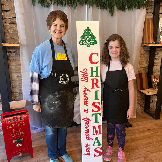 Two people in aprons standing next to a Christmas-themed porch sign with decorative elements.