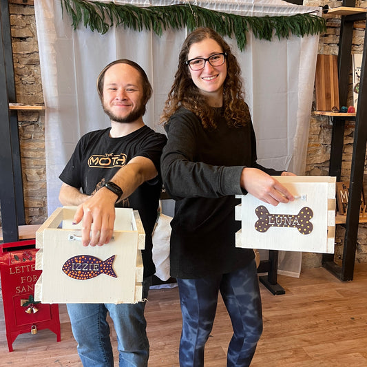 Two people standing on a wooden floor holding decorative pet toy boxes.
