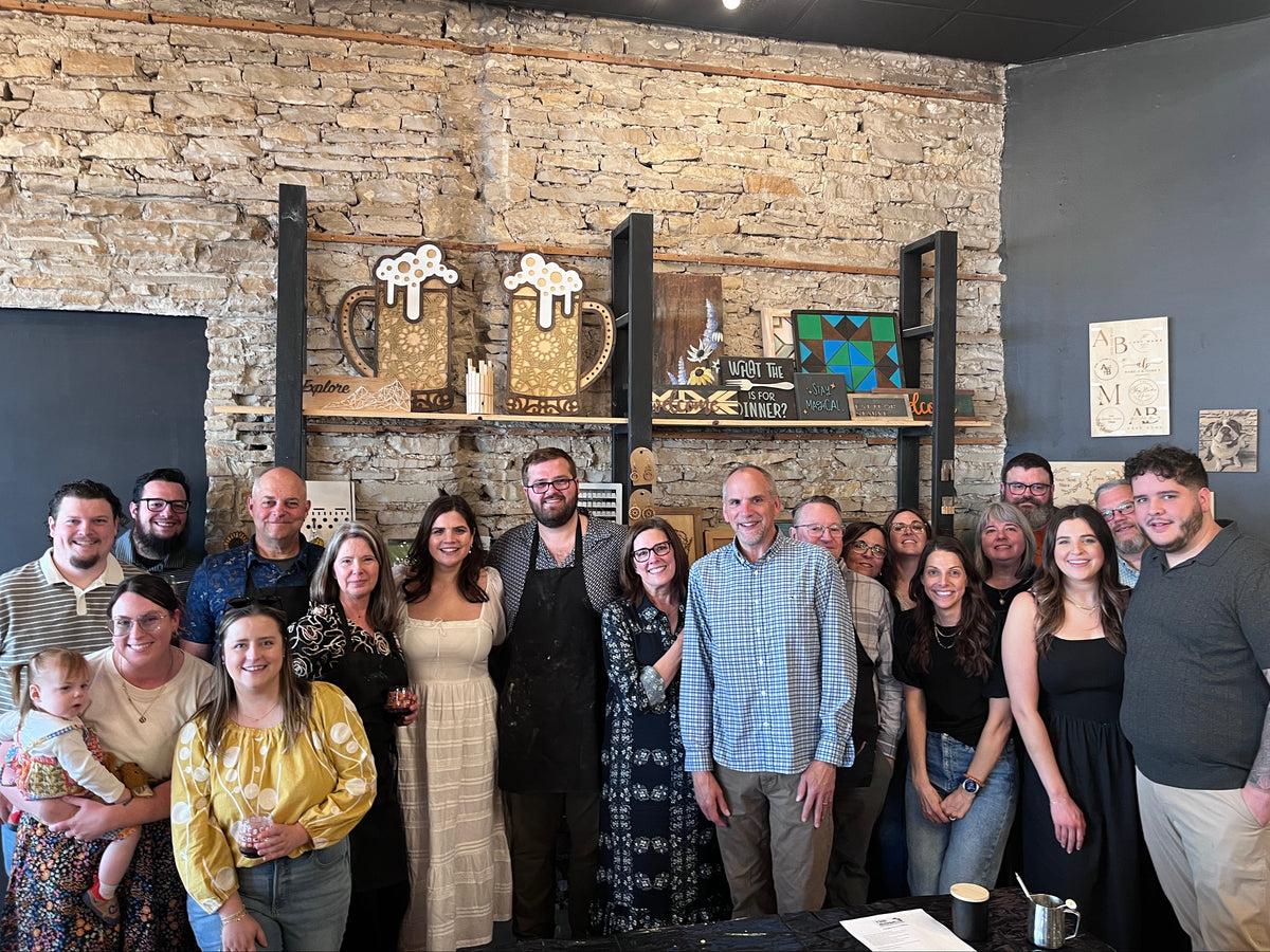 A group of people posing together in a rustic indoor space with a stone wall and shelving. They are guests at a wedding shower workshop.