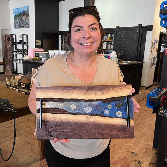 A smiling woman holds up an epoxy river tray with engraved live-edge wood, a blue floral inlay, and two black metal handles. The shop interior with tools and shelves is visible in the background.