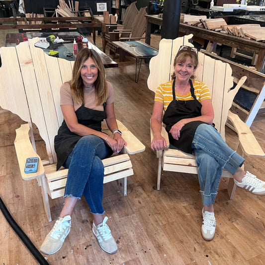 Two women wearing black aprons sit smiling in large unfinished wooden Adirondack chairs inside a woodworking workshop. The chairs are made from light natural wood planks and are shown in a real workshop environment with other furniture pieces and worktables in the background.