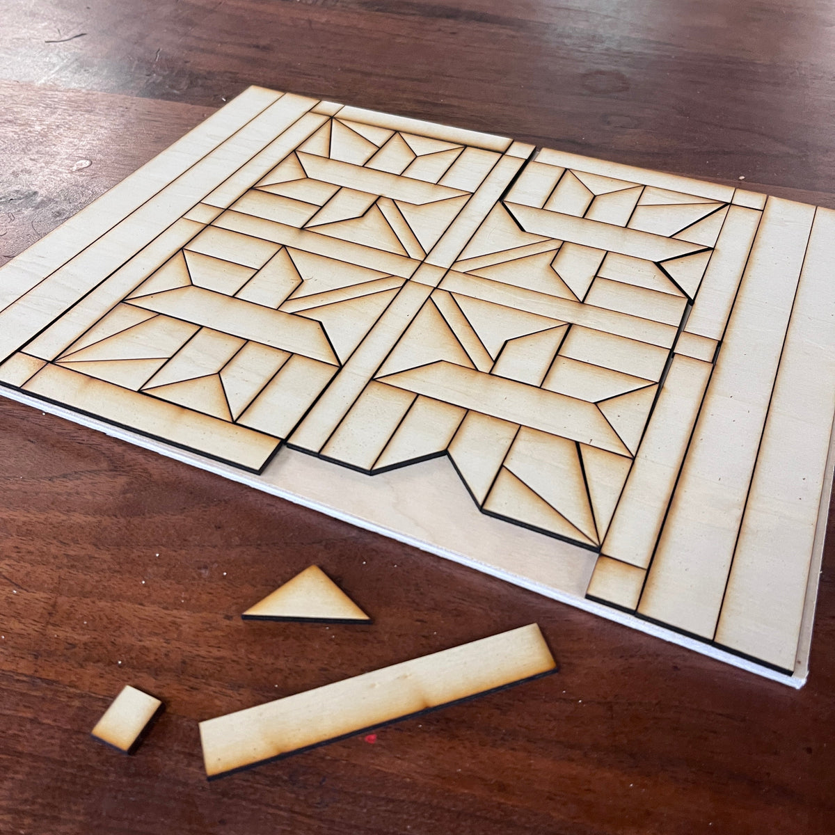 A wooden geometric design with laser-cut leaf patterned pieces, displayed on a dark wood tabletop with a few loose pieces in front.