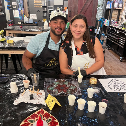 A man and a woman wearing black and white Mr. and Mrs. aprons sit behind a craft table covered in glass and epoxy materials, cups, and tools inside a workshop.