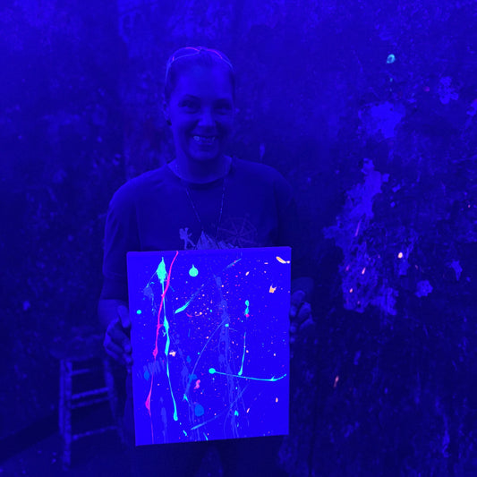 A person holding a canvas with fluorescent abstract splatter paint under blacklight, showing glowing neon blue, green, pink, and orange marks against a dark blue-lit background.