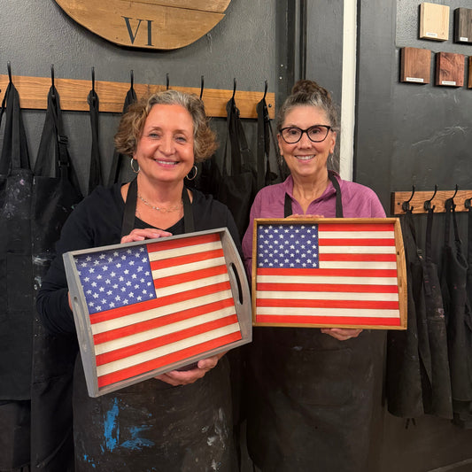 Two women wearing paint-splattered black aprons smile while holding wooden trays painted with the American flag in a workshop setting.