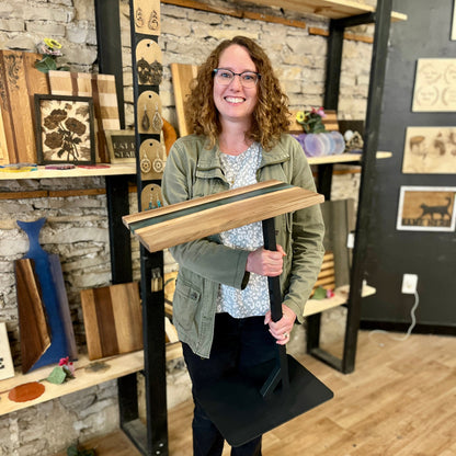 Woman holding a wooden epoxy river side table with live edge wood and a black metal base in a studio setting.