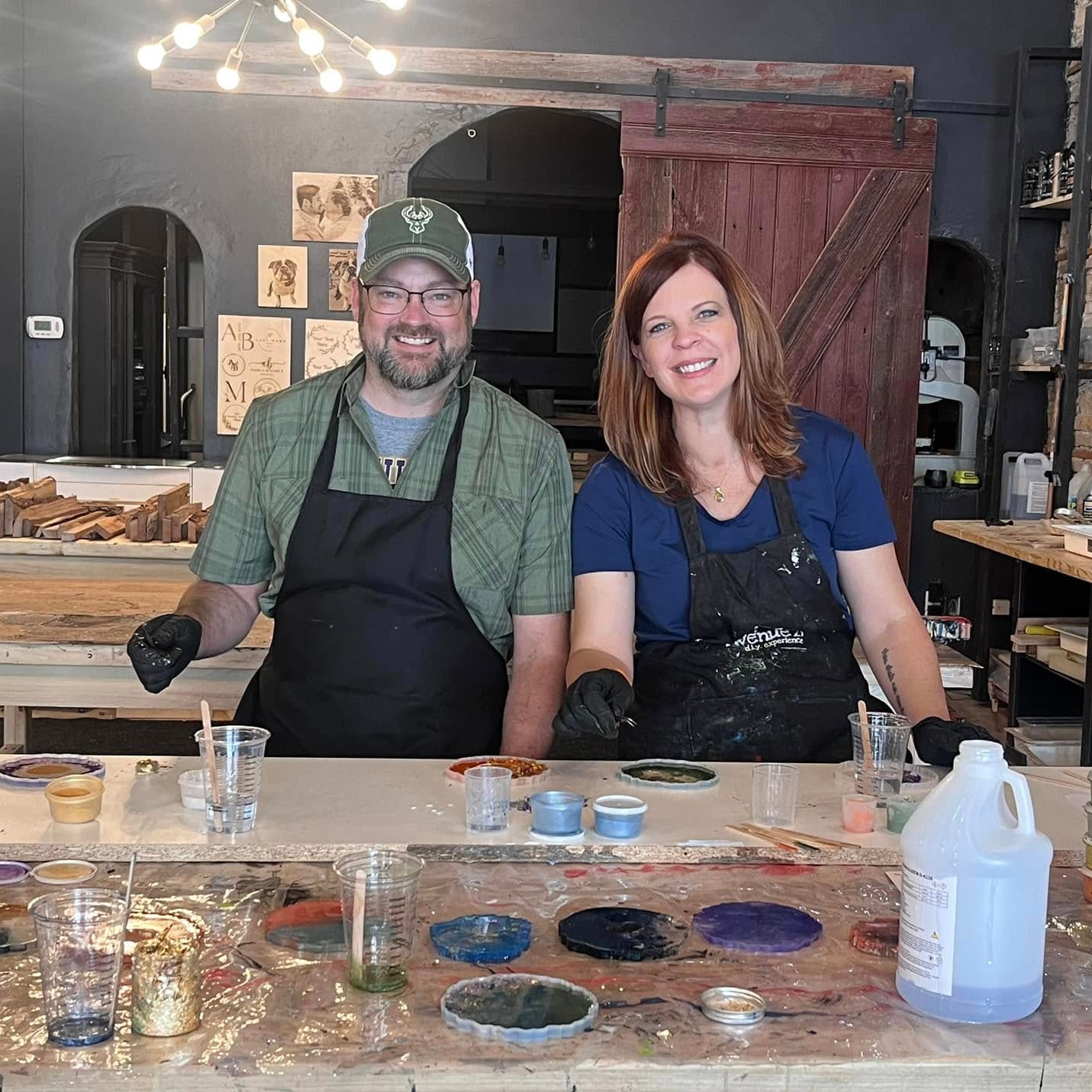 Two people in aprons at a table with epoxy coaster supplies in a workshop setting.