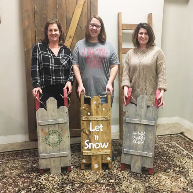 Three women holding wooden sleds with decorative messages indoors.
