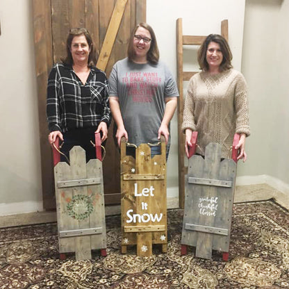 Three women holding wooden sleds with decorative messages indoors.