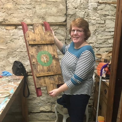 Woman holding a wooden sled with a green wreath against a stone wall.