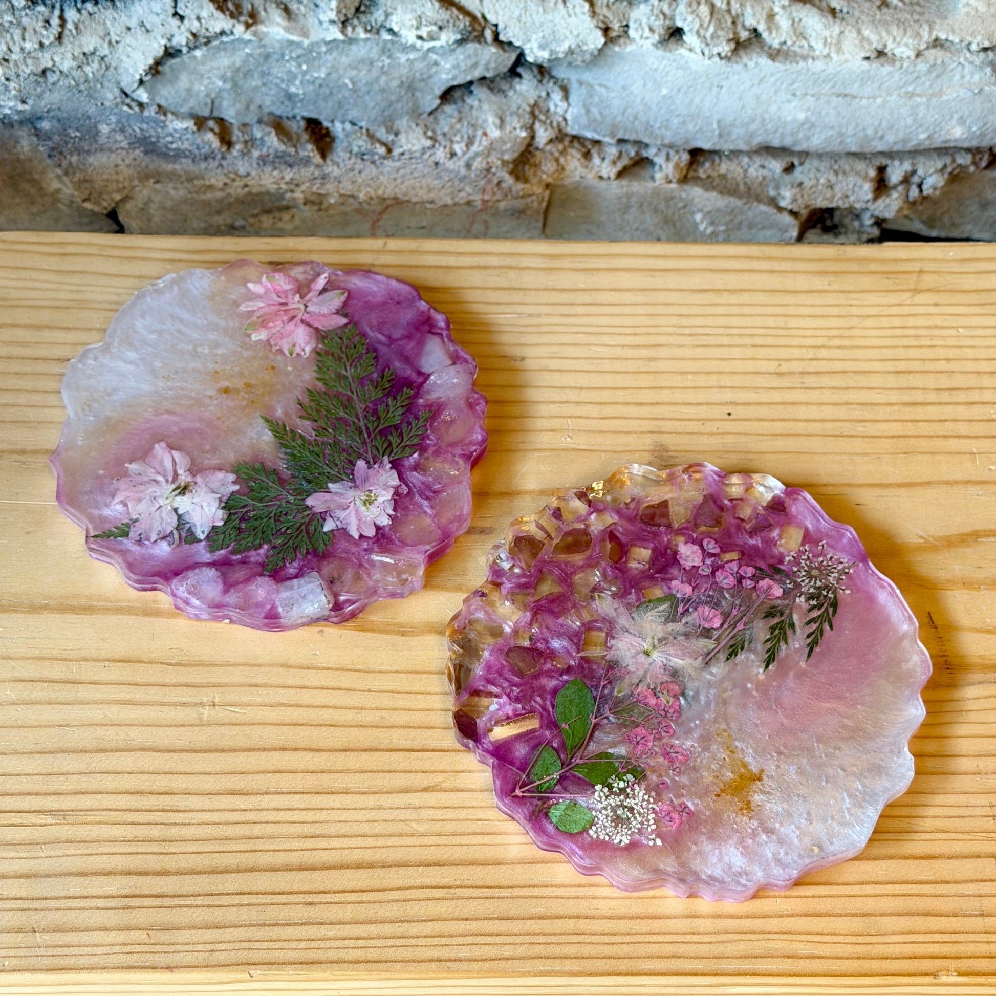 Two pink epoxy and glass crystal coasters with a floral embedded design on a wooden surface.