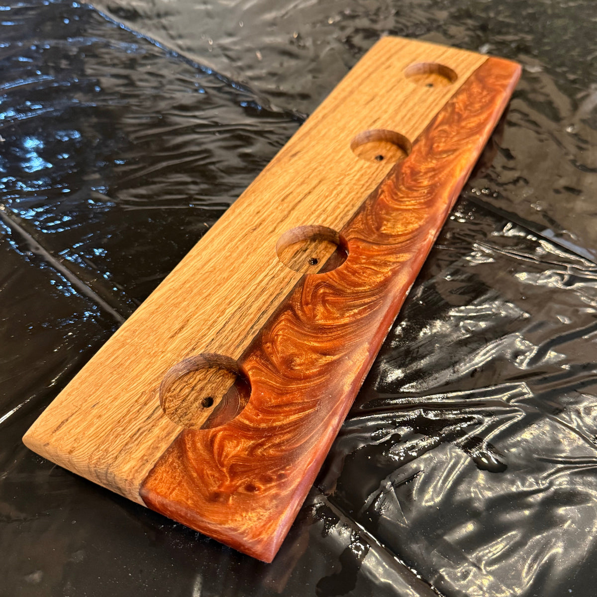 A rectangular wooden candle holder with three circular cutouts, featuring a natural wood grain pattern with reddish-brown swirls, resting on a black plastic sheet.