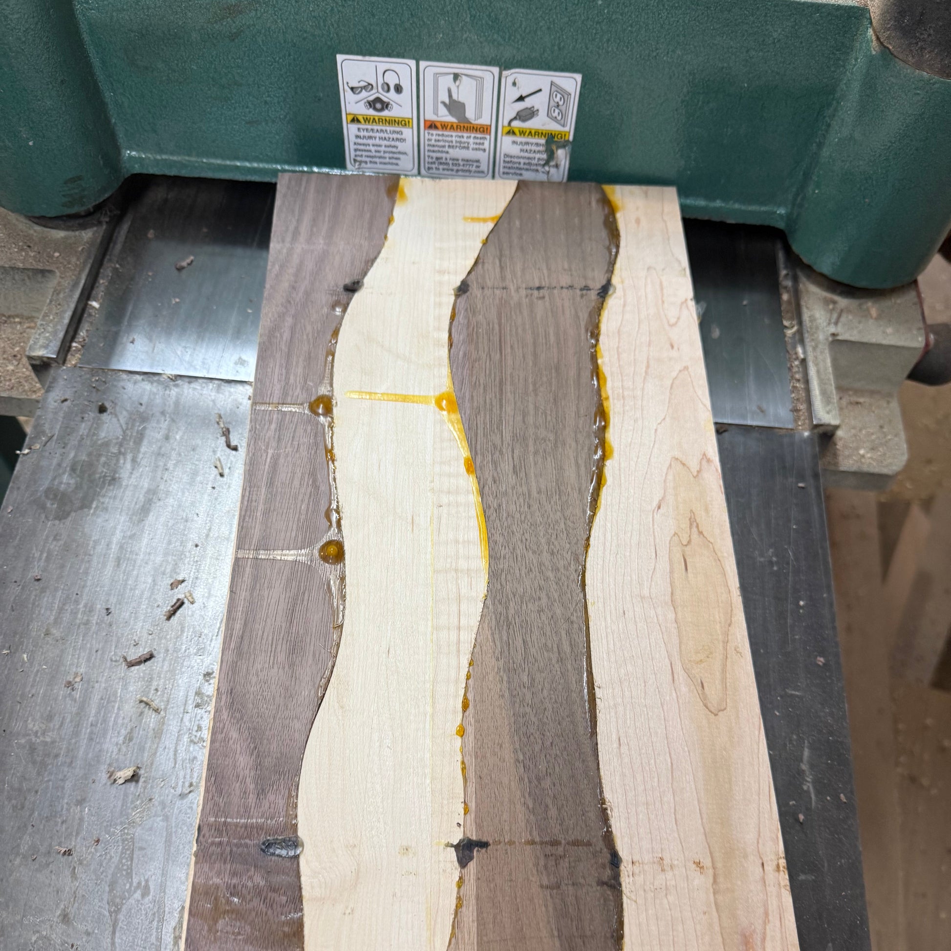 A rectangular wood slab with three vertical wavy bands of different wood tones and small amber resin pockets, resting on a metal workbench under a green planer machine.