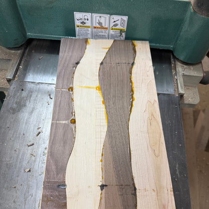 A rectangular wood slab with three vertical wavy bands of different wood tones and small amber resin pockets, resting on a metal workbench under a green planer machine.