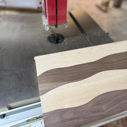 A close-up of a band saw in a workshop cutting a wood piece with a wavy, striped grain pattern of light and dark wood tones.