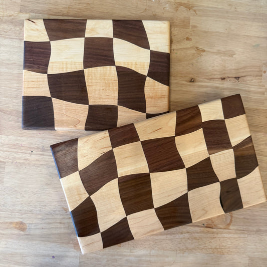 Two rectangular wooden cutting boards with a wavy checkerboard pattern in dark and light wood tones, displayed on a light wood table.