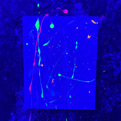 A canvas featuring neon splatters and drips in green, pink, orange, and blue in a splatter room with black lights.