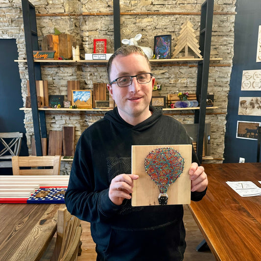 Man holding a wooden string art piece with a colorful hot air balloon design in a DIY studio room with shelves and tables.
