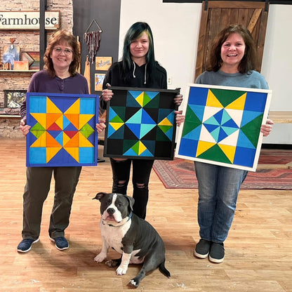 Four women holding colorful wooden quilt art with a dog in the foreground in a room with wooden floors and stone walls.