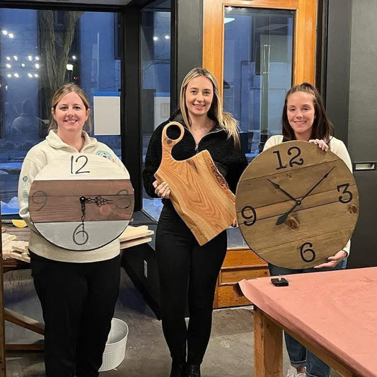 3 people holding wooden clocks and a charcuterie board in a workshop setting.