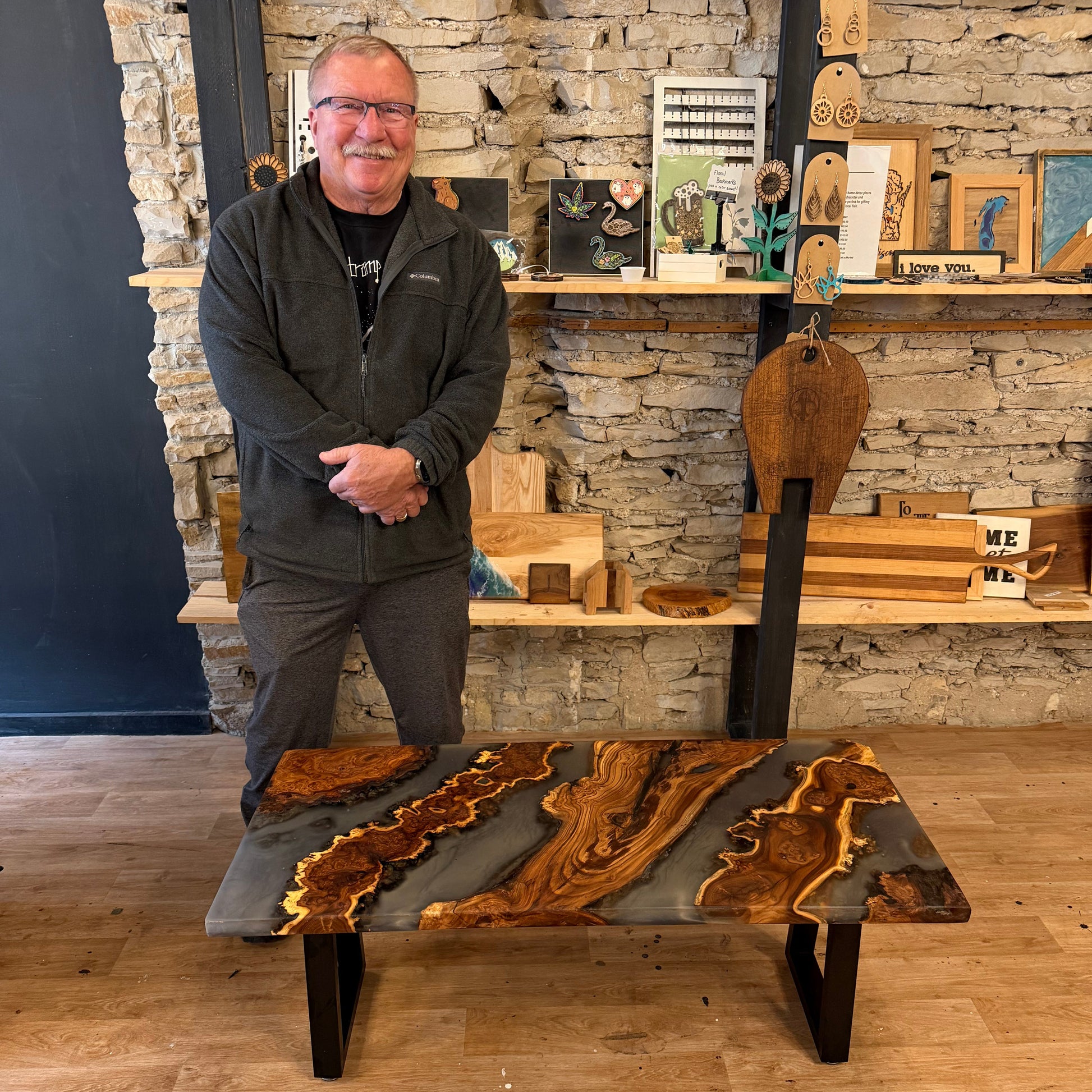 Man standing next to a wood and epoxy coffee table with in a rustic studio setting.
