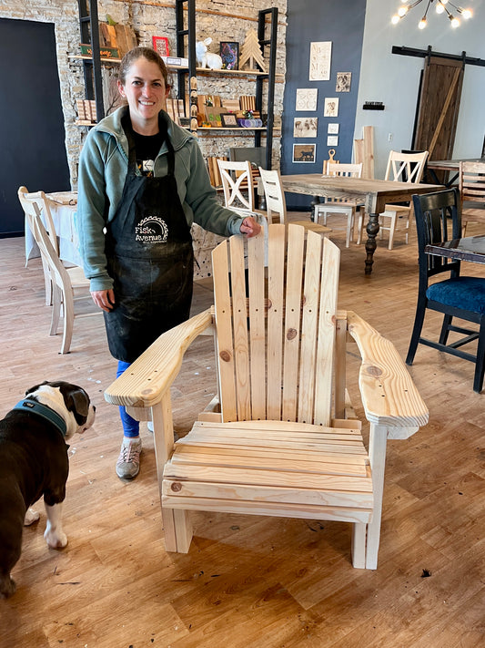 Woman in apron standing with a wooden Adirondack chair in a workshop with a dog.