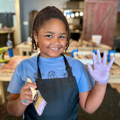 Child with an apron and a painted hand holding a paintbrush for a craft project in a workshop setting.