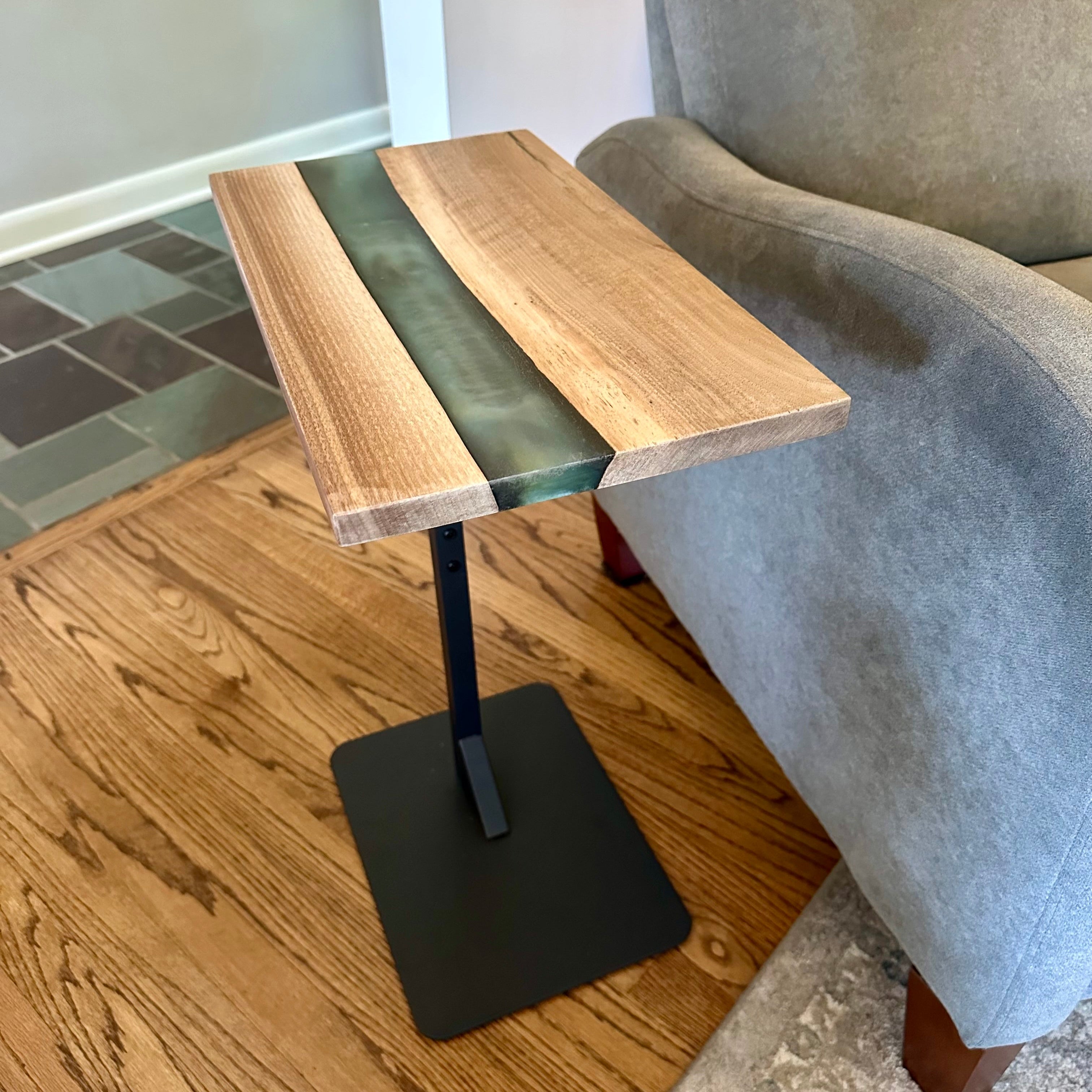 Small wood and epoxy table with black base next to a gray armchair on a wooden floor.